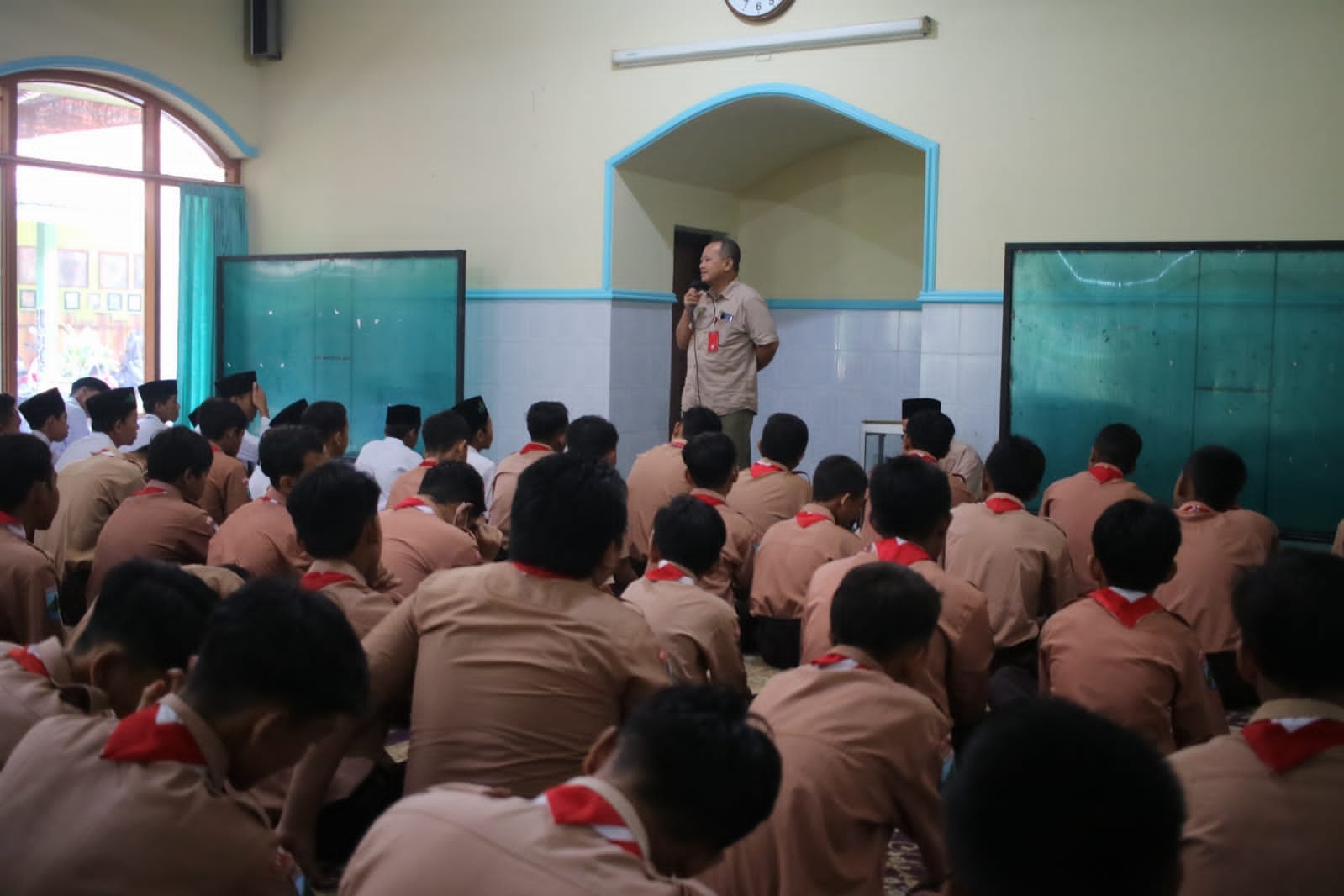 A warm and respectful Indonesian teacher greeting female and male middle school students warmly at the school gate under morning sunlight, surrounded by green tropical campus plants at SMPN 1 Gurah.