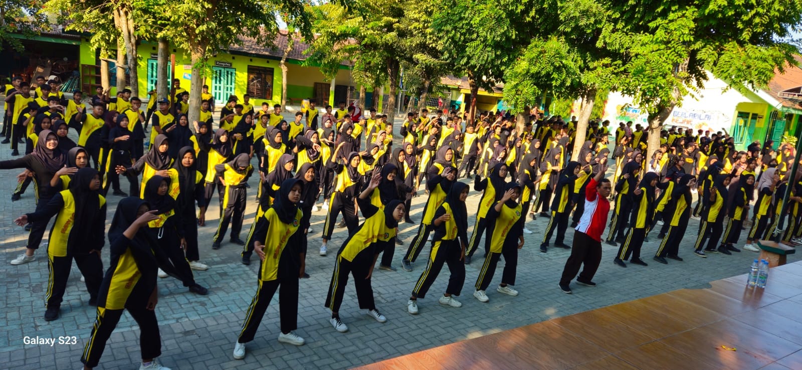 Energetic Indonesian middle school students in neat batik school uniforms gathered together outdoors in the green garden courtyard of SMPN 1 Gurah on a sunny Friday morning, smiling brightly with morning dew and lush tropical foliage.