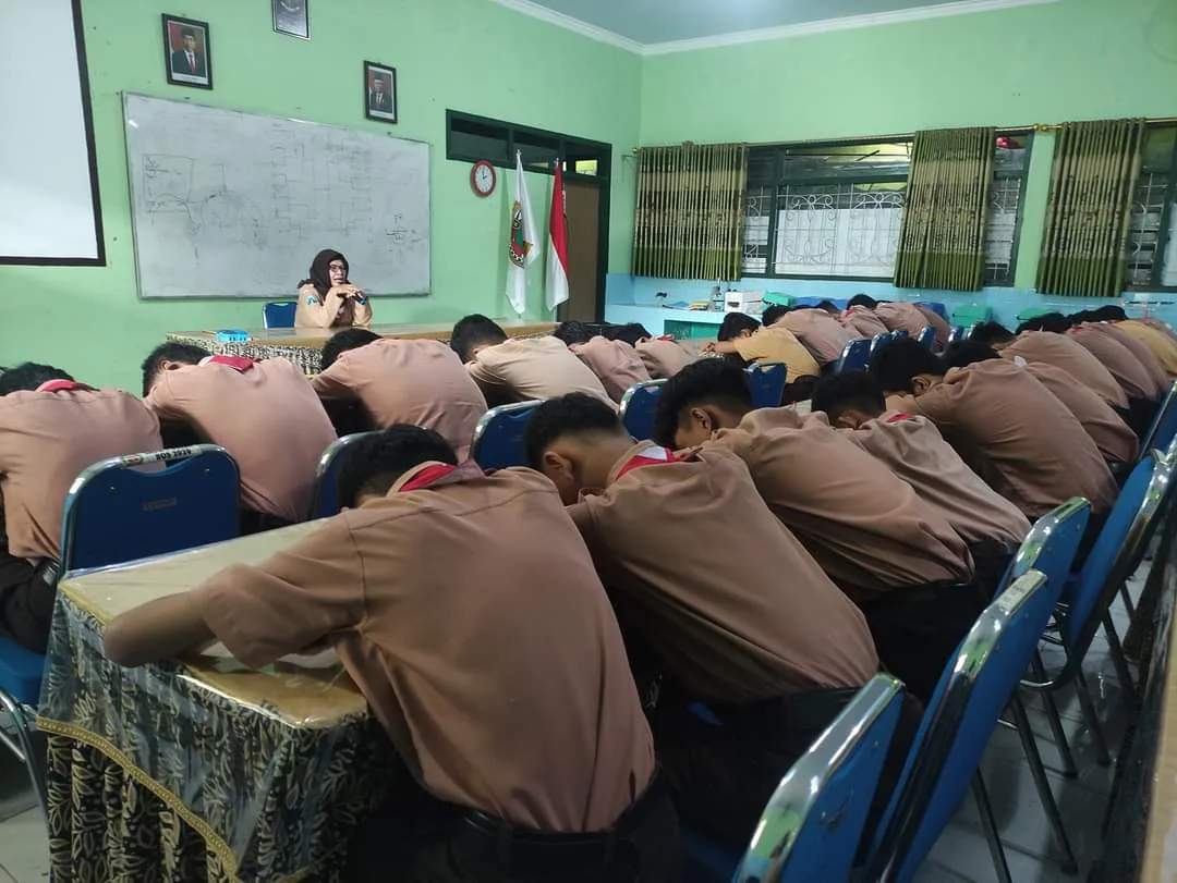 An Indonesian middle school homeroom teacher engaging in an intimate moral discussion with attentive students sitting inside a clean bright classroom with wooden desks and open windows.